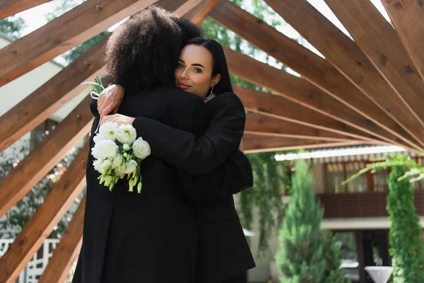 Лесбіянка з весільним букетом обіймає афроамериканську дівчину в парку. — стокове фото