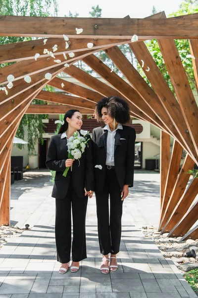 Весела міжрасова лесбійська пара з весільним букетом, що стоїть під пелюстками троянд в парку — стокове фото