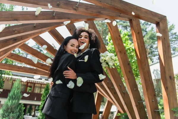 Fröhliches multiethnisches lesbisches Paar in Anzügen, das sich bei der Hochzeit im Park in der Nähe von Blütenblättern umarmt — Stockfoto