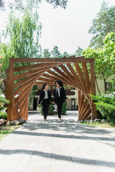 Sonriente pareja de lesbianas multiétnicas en trajes con ramo de bodas caminando en el parque - foto de stock