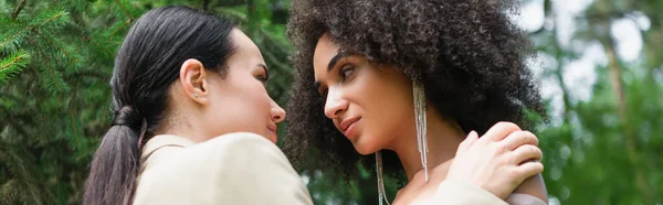 Low angle view of lesbian woman in suit hugging african american girlfriend in park, banner — Stock Photo
