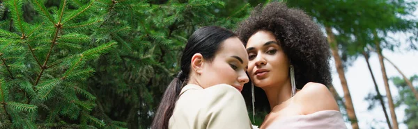 Vista de ángulo bajo de la mujer lesbiana afroamericana de pie cerca de su novia en traje en el parque, pancarta - foto de stock