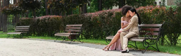 Interracial lesbian couple in suit and dress sitting on bench in park, banner — Stock Photo