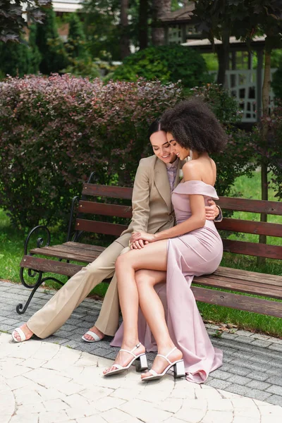 Sorridente interrazziale lesbica coppia abbracciare su panchina — Foto stock