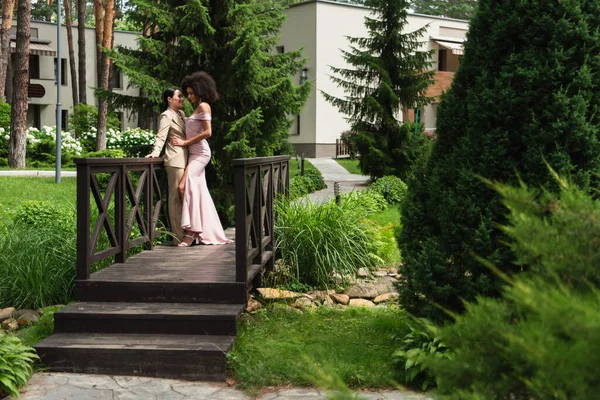 Cheerful interracial lesbian couple in formal wear hugging on bridge — Stock Photo