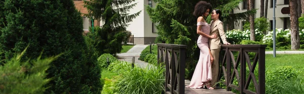 Afroamerikanerin im Kleid umarmt lächelnde Freundin im Anzug auf Brücke, Banner — Stockfoto