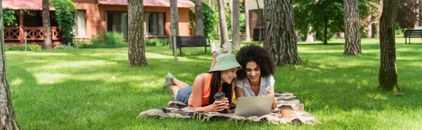 Alegre pareja lesbiana interracial usando teléfono inteligente cerca de la computadora portátil y café en el parque, pancarta - foto de stock