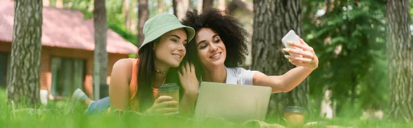 Sonriente pareja lesbiana interracial con café tomando selfie cerca del portátil en el parque, pancarta - foto de stock