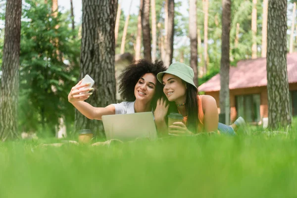 African american woman taking selfie near girlfriend with coffee and laptop in park — Stock Photo