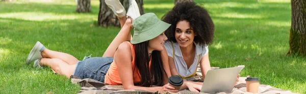 Sonriente pareja lesbiana interracial utilizando dispositivos cerca de vasos de papel en manta en el parque, pancarta - foto de stock