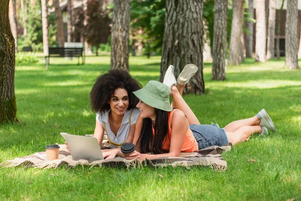 Cheerful interracial same sex couple with coffee to go using devices on blanket in park — Stock Photo