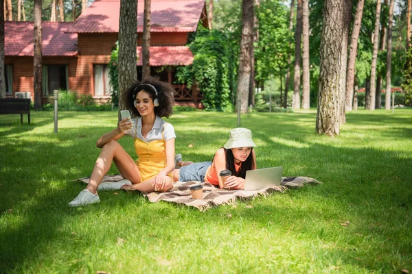 African american woman in headphones using smartphone near girlfriend with coffee and laptop on lawn — Stock Photo