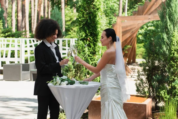 Alegre pareja interracial tintineo con champán durante la boda al aire libre - foto de stock