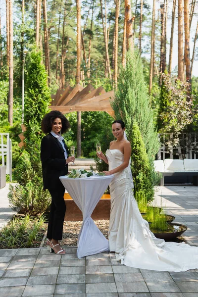 Sonriente pareja lesbiana interracial con champán de pie cerca de ramo durante la boda al aire libre - foto de stock