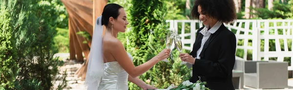 Positivo interracial lesbianas pareja tintineo con champán durante la boda al aire libre, bandera - foto de stock