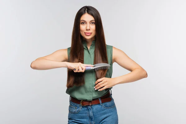 Lächelnde junge Frau beim Bürsten glänzendes Haar isoliert auf grau — Stockfoto