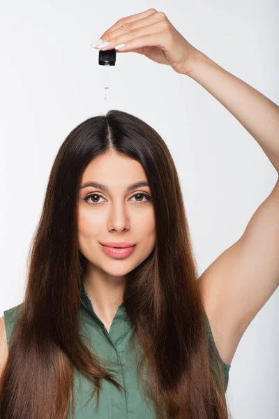 Junge Frau mit glänzenden Haaren, die Behandlungsöl auf den Kopf aufträgt, isoliert auf grau — Stockfoto