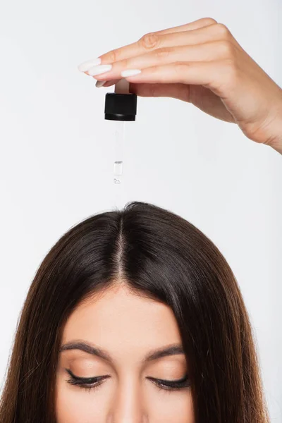 Обрезанный вид молодой женщины с блестящими волосами, применяющей лечебное масло на верхней части головы изолированы на сером — стоковое фото