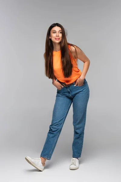 Volle Länge der glücklichen jungen Frau in Jeans posiert mit Händen in Taschen auf grau — Stockfoto