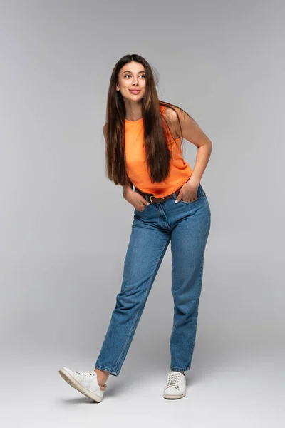 Pleine longueur de jeune femme heureuse en jeans en denim posant avec les mains dans les poches sur gris — Photo de stock