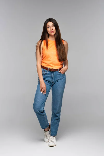 Full length of happy young woman in denim jeans posing with hand in pocket on grey — Stock Photo