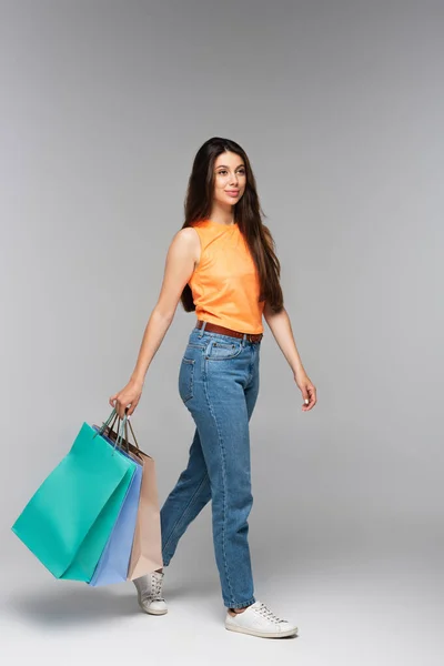 Longitud completa de morena mujer joven sosteniendo bolsas de compras y caminando sobre gris - foto de stock