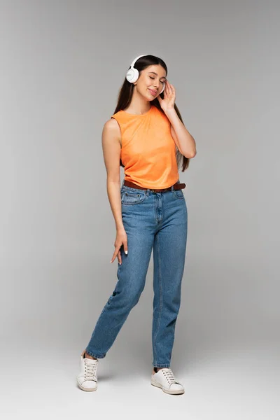 Longitud completa de la mujer sonriente en jeans y auriculares inalámbricos escuchando música en gris - foto de stock