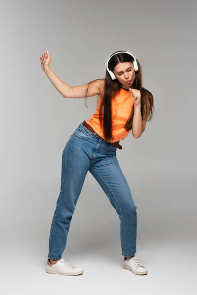 Full length of brunette woman in denim jeans and wireless headphones listening music while dancing on grey — Stock Photo