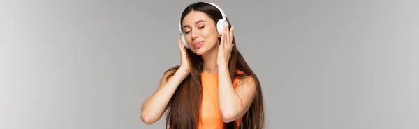 Pleased woman in denim jeans and wireless headphones listening music and smiling isolated on grey, banner — Stock Photo