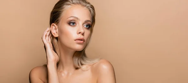 Bonita mujer con maquillaje en la cara perfecta tocando el pelo mientras mira hacia otro lado aislado en beige, pancarta — Stock Photo