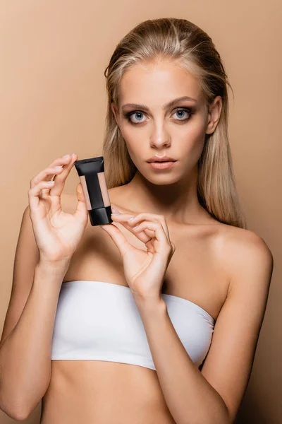 Blonde woman with grey eyes showing tube of makeup foundation isolated on beige — Stock Photo
