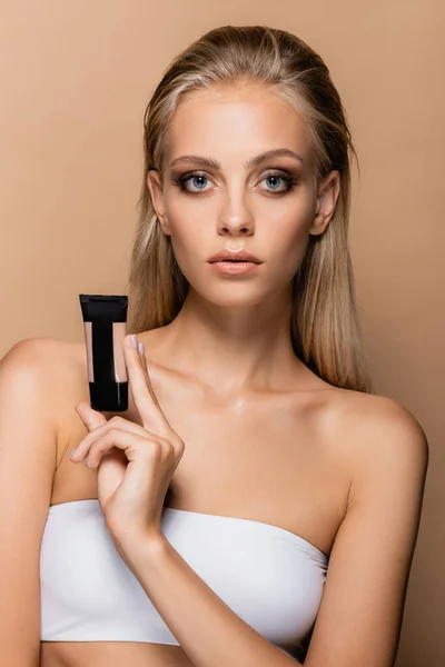 Pretty blonde woman with grey eyes showing tube with face foundation isolated on beige — Stock Photo