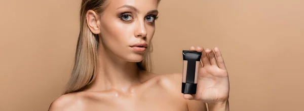 Young blonde woman showing tube with face foundation isolated on beige, banner — Stock Photo