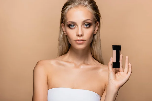 Pretty woman with grey eyes and blonde hair showing tube of makeup foundation isolated on beige — Stock Photo