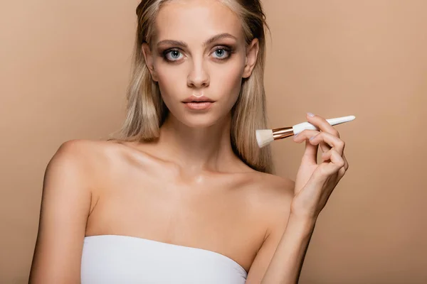 Blonde woman with naked shoulders posing with cosmetic brush isolated on beige — Stock Photo