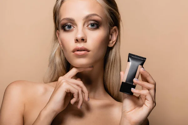 Woman with blonde hair and bare shoulders holding tube of makeup foundation isolated on beige — Stock Photo
