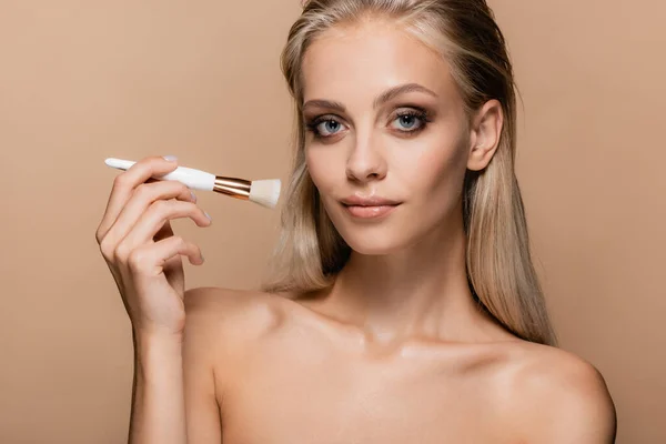 Blonde woman with cosmetic brush smiling at camera isolated on beige — Stock Photo
