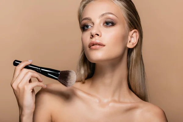 Blonde woman with makeup on perfect face holding soft cosmetic brush isolated on beige — Stock Photo