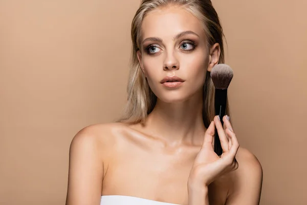 Jolie femme aux cheveux blonds et maquillage posant avec une grande brosse cosmétique isolée sur beige — Photo de stock