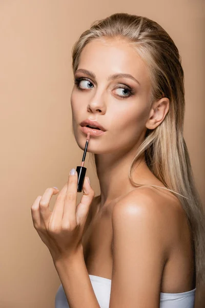 Blonde woman with naked shoulders applying pink lip gloss isolated on beige — Stock Photo