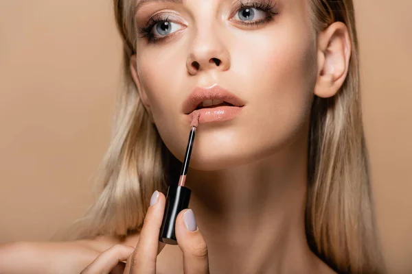 Young woman with perfect face applying pastel pink lip gloss isolated on beige — Stock Photo