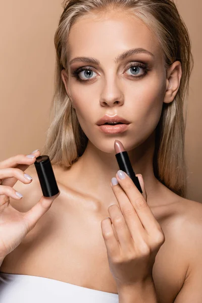 Blonde woman with perfect skin and grey eyes holding lipstick isolated on beige — Stock Photo