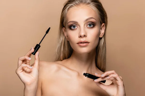 Pretty woman with blonde hair and naked shoulders holding black mascara isolated on beige — Stock Photo