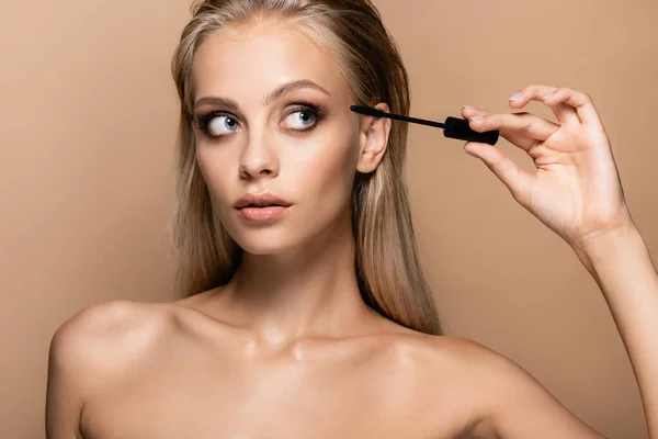 Young woman with bare shoulders looking at black mascara isolated on beige — Stock Photo