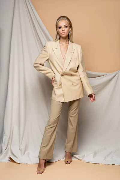 Mujer rubia en traje elegante posando con la mano en la cadera cerca de fondo beige drapeado — Stock Photo