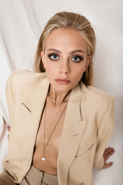 High angle view of young blonde woman in beige unbuttoned blazer sitting on fabric and looking at camera — Stock Photo
