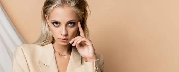 Mujer rubia con maquillaje sosteniendo la mano cerca de la cara sobre fondo beige drapeado, pancarta - foto de stock