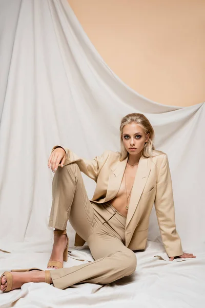 Mujer sensual en traje de moda mirando a la cámara mientras está sentado sobre fondo beige con cortinas - foto de stock