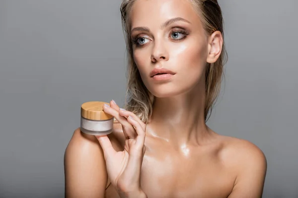 Mulher loira com pele molhada segurando creme cosmético perto do ombro nu isolado no cinza — Fotografia de Stock