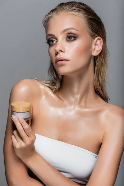 Wet woman with blonde hair holding cosmetic cream isolated on grey — Stock Photo